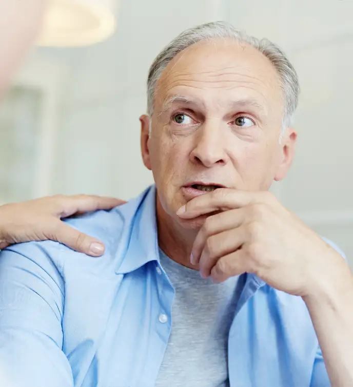 TraumaSolutions An older man with gray hair wearing a blue shirt looks thoughtful, resting his hand on his chin, while another person—possibly a clinician—places a reassuring hand on his shoulder, illustrating support often seen in Brainspotting sessions.