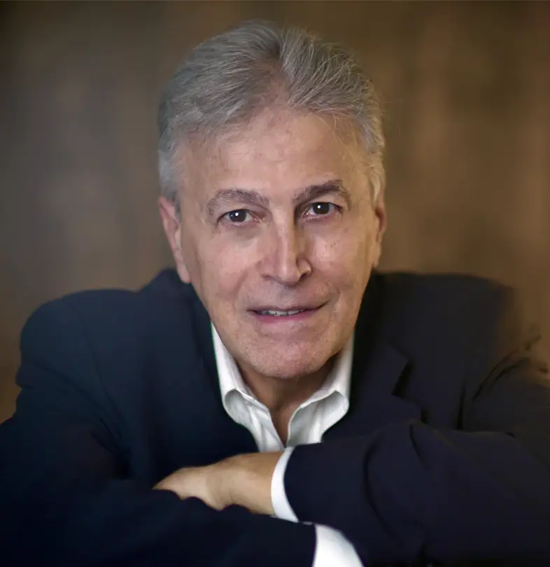 TraumaSolutions Dr. David Grand with gray hair, wearing a dark suit jacket and white shirt, sits with his arms crossed, looking at the camera against a brown background—an ideal image for promoting Brainspotting masterclass for clinicians.