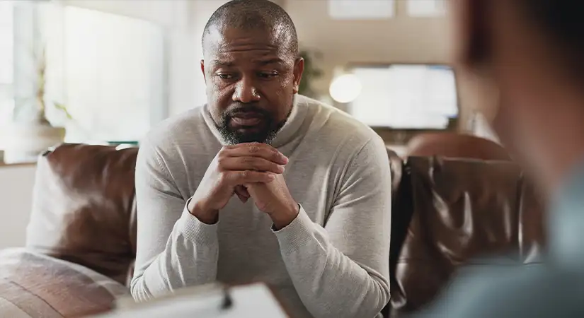 TraumaSolutions A man sits on a couch with his hands clasped, appearing thoughtful or concerned, while another person—perhaps during a Brainspotting masterclass for clinicians—sits across from him holding a clipboard.