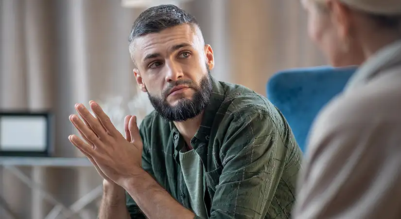 TraumaSolutions A bearded man sits indoors, looking attentively at another person during a conversation about Brainspotting, his hands clasped together as if engaged in a masterclass for clinicians.