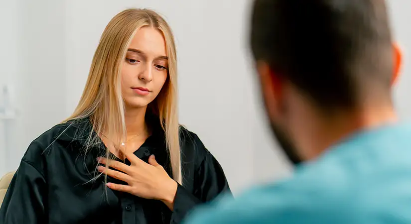 TraumaSolutions A woman with long blonde hair sits across from a man, touching her chest with one hand, appearing to express emotion during a Brainspotting masterclass conversation.