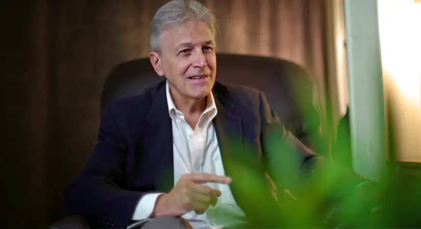 TraumaSolutions An older man in a suit jacket sits in an armchair, gesturing with his hand, as if leading a masterclass for clinicians, with a green plant in the foreground.
