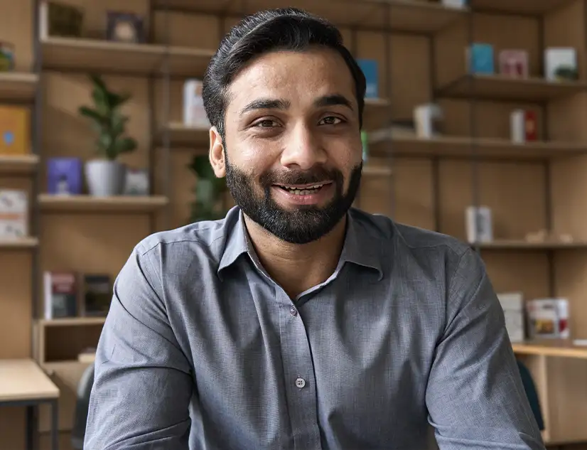 TraumaSolutions A man with a beard in a gray shirt sits at a desk, smiling, with shelves of books and plants behind him—ready to lead a masterclass for clinicians on Brainspotting and working with what arises.