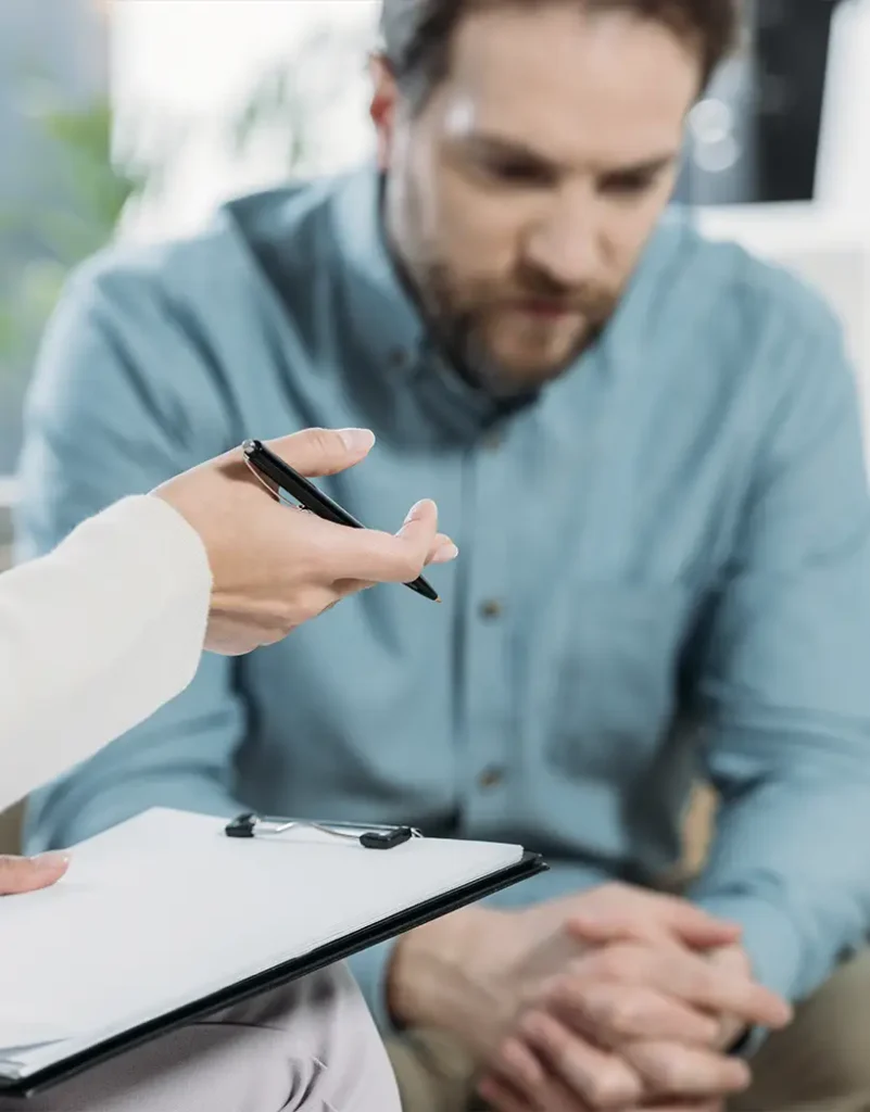 TraumaSolutions A man sits with his hands clasped, looking down, while another person holds a clipboard and pen, suggesting a Brainspotting therapy session focused on trauma and deep processing.