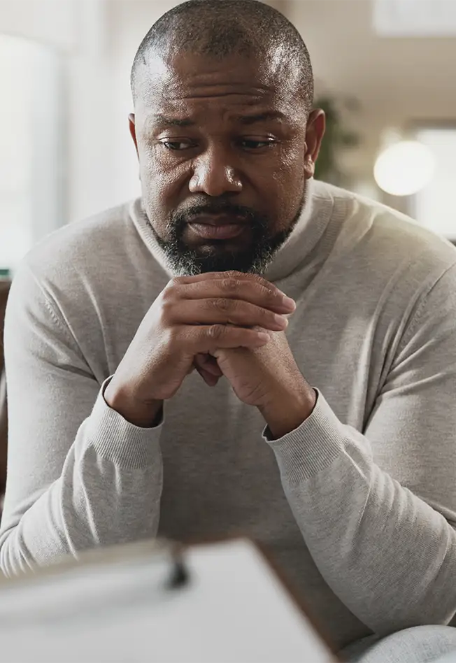 TraumaSolutions A man in a light gray turtleneck sits with his hands clasped, looking down with a serious expression, as if engaged in deep processing or reflecting on trauma.