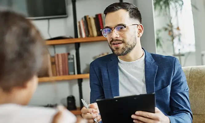 TraumaSolutions A man in glasses and a blue blazer holds a clipboard and pen, discussing deep processing or Brainspotting with another person in a living room or office setting.