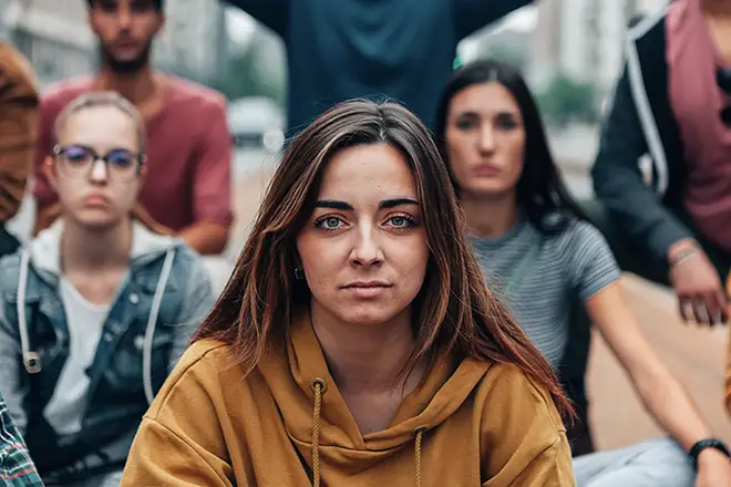 TraumaSolutions A group of young adults sitting outdoors, with a woman in a yellow hoodie in the foreground looking directly at the camera, capturing a moment of deep processing and connection.