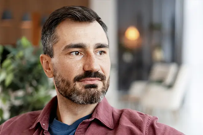 TraumaSolutions A man with short dark hair and a beard, wearing a maroon shirt over a blue t-shirt, looks to his left in a softly lit indoor setting—suggesting deep processing or reflection often seen in trauma-focused therapies like Brainspotting for clinicians.