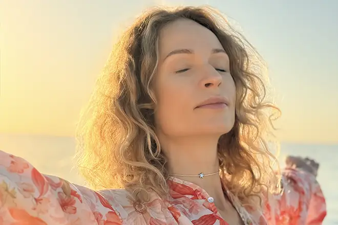 TraumaSolutions A woman with curly blonde hair stands with eyes closed and arms outstretched, wearing a floral top, in soft sunlight, as if experiencing deep processing—a moment of release often found in Brainspotting therapy for trauma.