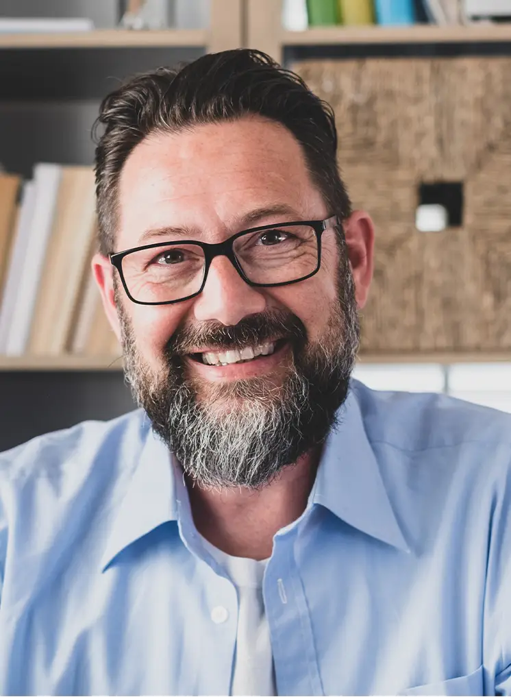 TraumaSolutions A man with glasses and a beard, wearing a light blue shirt, smiles while sitting indoors with shelves and books in the background, suggesting a setting ideal for deep processing techniques like Brainspotting to address trauma.
