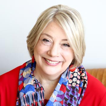 TraumaSolutions Smiling older woman with short blonde hair, wearing a red top and a colorful patterned scarf, seated in front of a light background—embodying hope and resilience in the journey of healing from complex trauma.