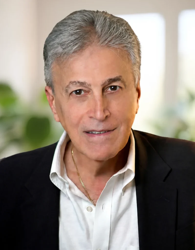 TraumaSolutions A middle-aged man with gray hair, wearing a dark blazer, white shirt, and gold necklace, stands indoors with a blurred background—ready to lead a Brainspotting masterclass for clinicians.