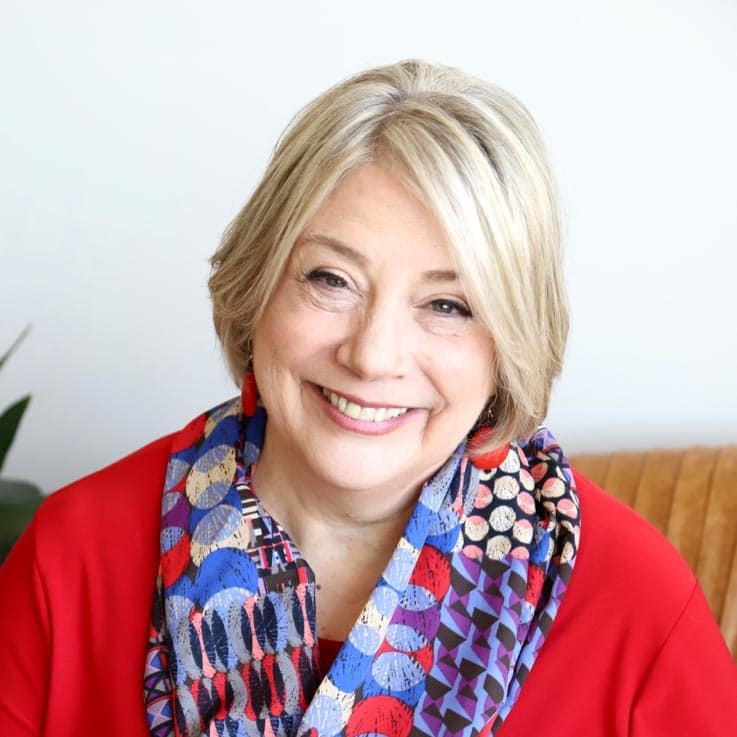 TraumaSolutions A smiling older woman with blonde hair wears a red top and a colorful patterned scarf, sitting indoors against a neutral background.