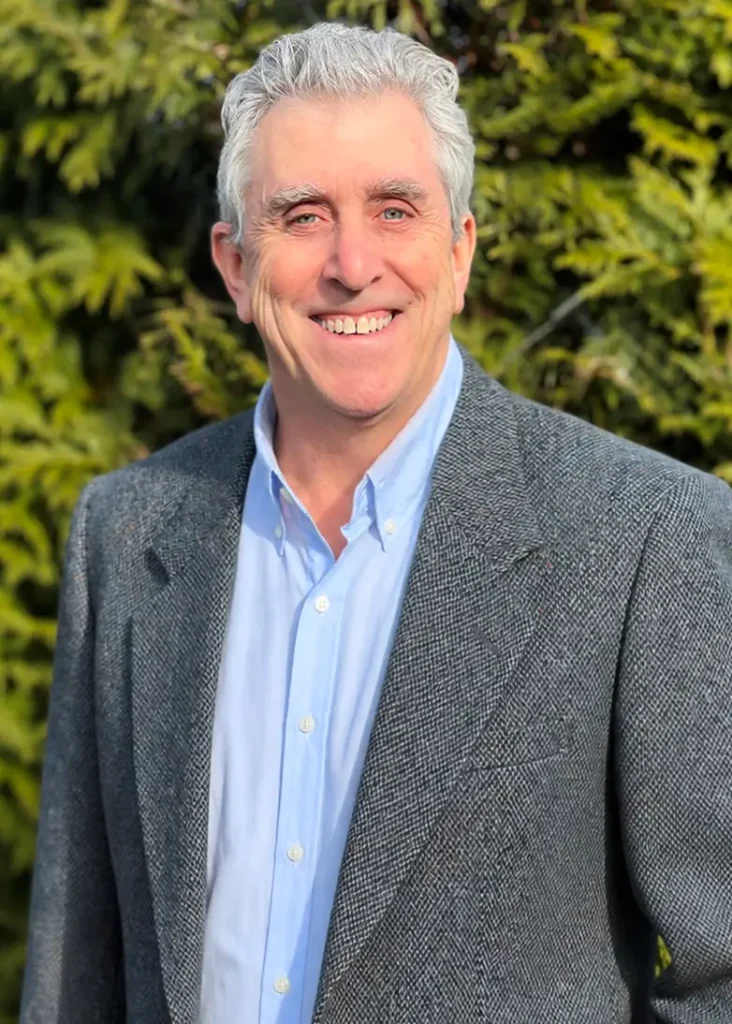 TraumaSolutions A man with gray hair wearing a gray blazer and light blue shirt stands outdoors in front of green foliage, smiling at the camera—radiating the calm confidence often found through deeper healing experiences such as EFT or trauma therapy.