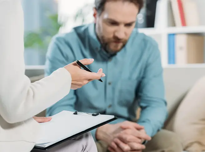TraumaSolutions A person holding a clipboard and pen speaks to a man sitting with his hands clasped, looking down, in what appears to be a trauma therapy session focused on deeper healing.