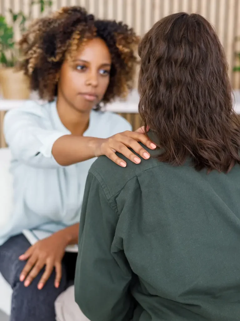 TraumaSolutions A woman places her hand on another person’s shoulder in a comforting gesture during a conversation indoors, offering support that can be essential on the journey toward deeper healing through trauma therapy.
