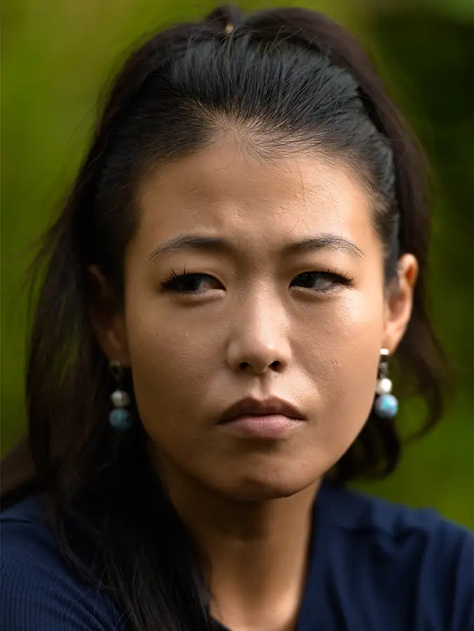 TraumaSolutions A woman with long dark hair and earrings looks to the side with a serious expression, set against a blurred green background—capturing a quiet moment that reflects the journey of deeper healing and trauma therapy.