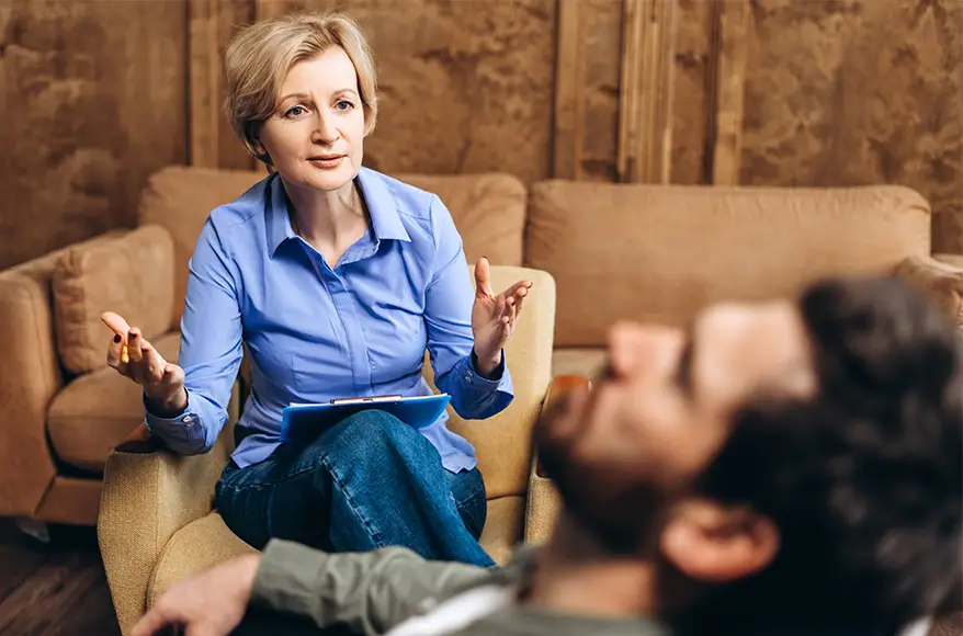 TraumaSolutions A woman with a clipboard listens to a man lying on a couch, suggesting an EFT-based therapy or trauma therapy session in a comfortable room focused on deeper healing.