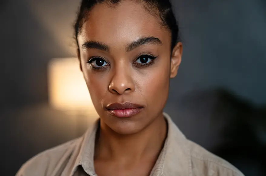 TraumaSolutions A woman with dark hair tied back looks directly at the camera with a neutral expression. She wears a beige shirt and stands indoors, the blurred lamp in the background suggesting a calming environment, ideal for trauma therapy or deeper healing.