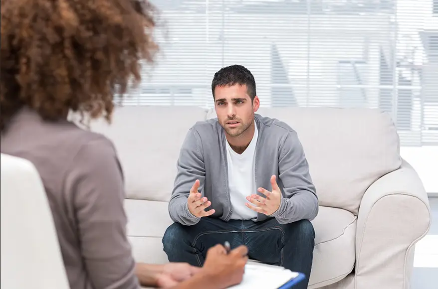 TraumaSolutions A man sits on a couch talking to a woman holding a notepad, suggesting a trauma therapy or counseling session in a bright office setting focused on deeper healing.