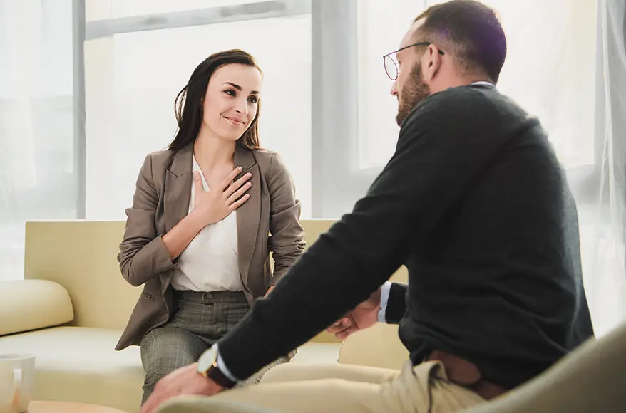 TraumaSolutions Two people sit on a couch having a conversation. The woman smiles with her hand on her chest, a gesture often seen during EFT sessions, while the man faces her and gestures with his hand, fostering an environment for deeper healing.