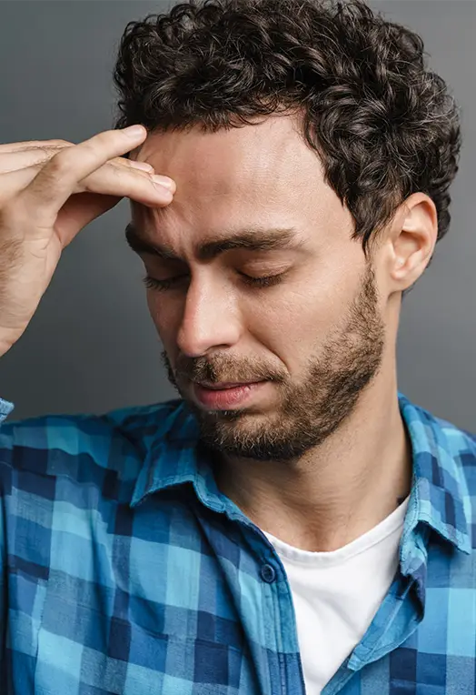 TraumaSolutions Man with curly hair and beard wearing a blue plaid shirt touches his forehead with eyes closed, appearing thoughtful or stressed—possibly reflecting on his journey through trauma therapy and seeking deeper healing.