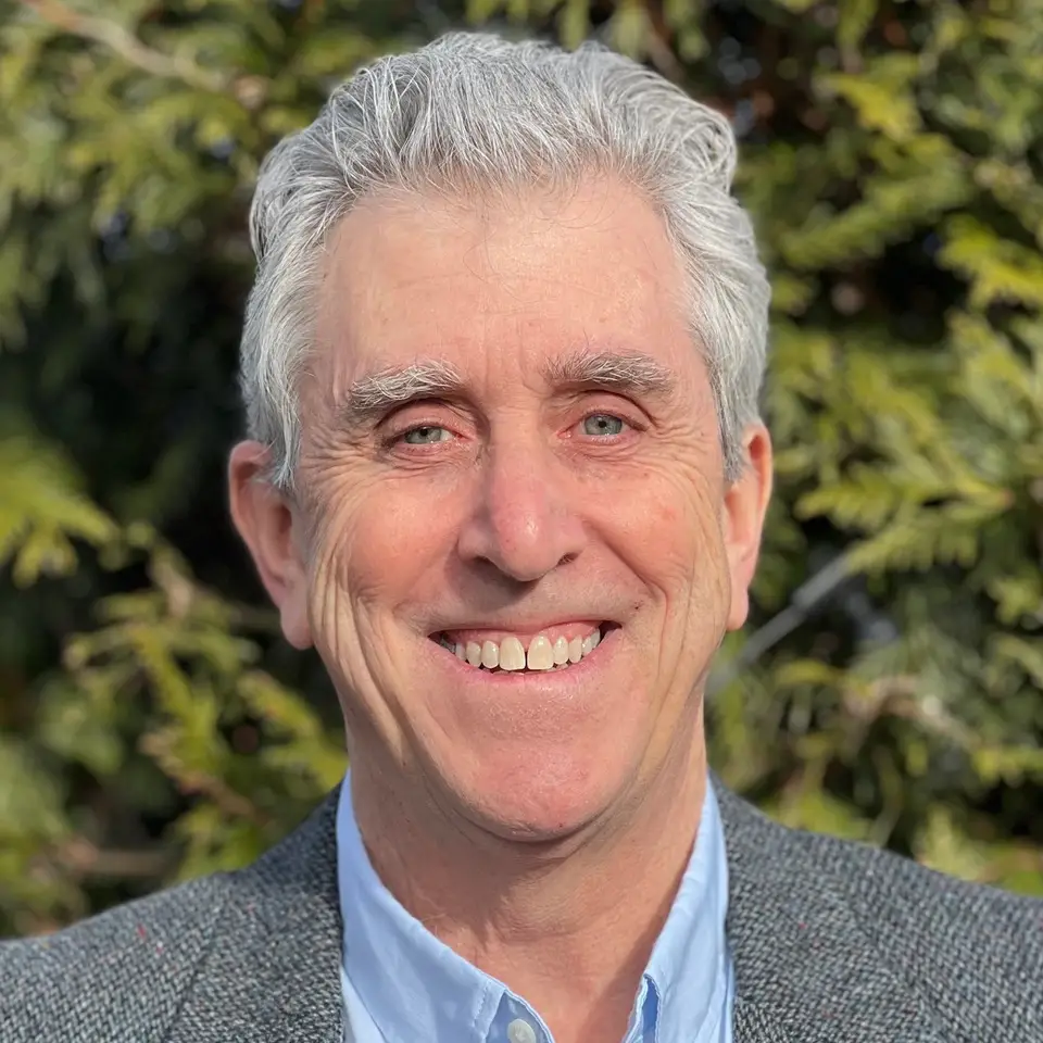 TraumaSolutions A man with gray hair and blue eyes smiles at the camera, wearing a gray blazer and blue shirt, standing outdoors in front of green foliage—a warm presence suggesting the calm assurance often found in those experienced with trauma therapy and dedicated to deeper healing.