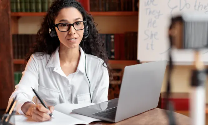 TraumaSolutions Woman wearing glasses and headphones sits at a desk with a laptop, writing in a notebook, with bookshelves and a whiteboard in the background.