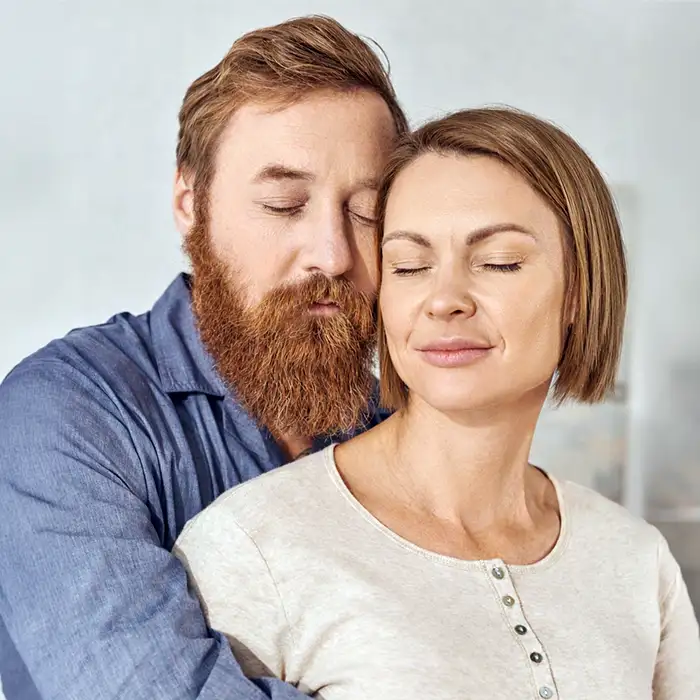 TraumaSolutions A man with a beard embraces a woman from behind. Both have their eyes closed and appear relaxed, exuding a sense of belonging in the softly lit indoor setting.