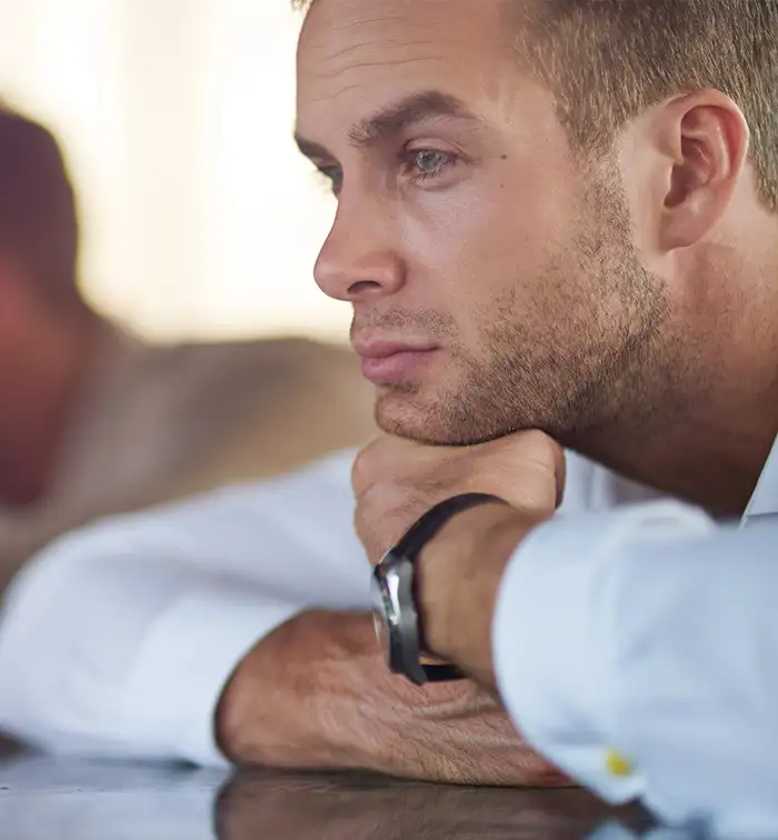 TraumaSolutions A man with short hair and stubble, wearing a white shirt and wristwatch, leans his chin on his hand and looks thoughtfully into the distance, reflecting on belonging and secure attachment.