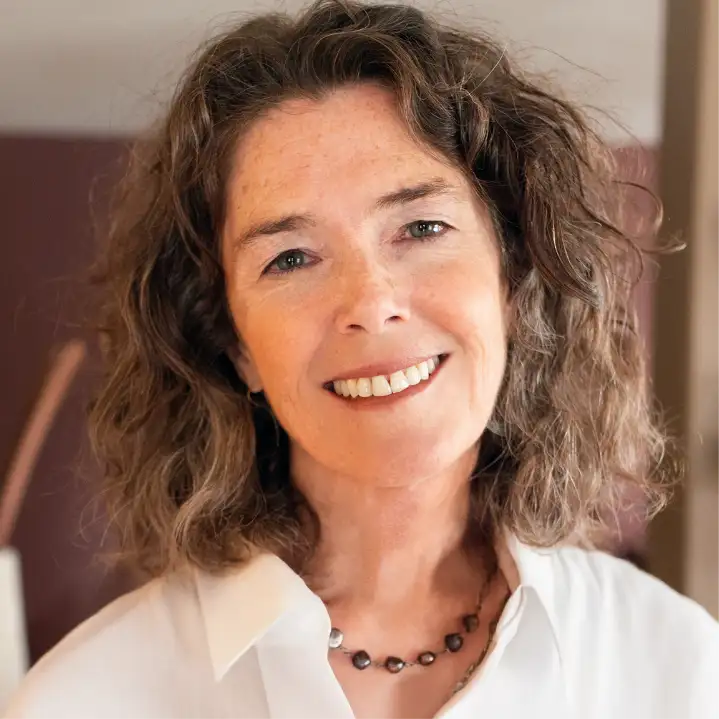 TraumaSolutions A woman with curly brown hair, wearing a white shirt and beaded necklace, smiles at the camera against a blurred indoor background, radiating a sense of belonging.