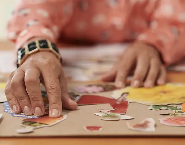 TraumaSolutions Close-up of a person arranging colorful paper cutouts on a table, wearing a patterned long-sleeve shirt and a black wristwatch—an act reflecting longing for belonging through creative expression.