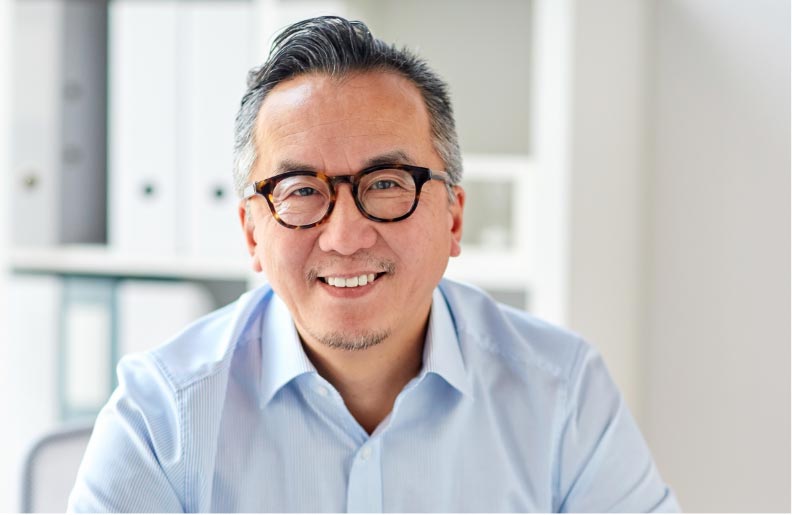 TraumaSolutions A man with glasses and gray hair, wearing a light blue shirt, smiles while sitting in a bright office space, reflecting on healing from trauma and loss through IFS.