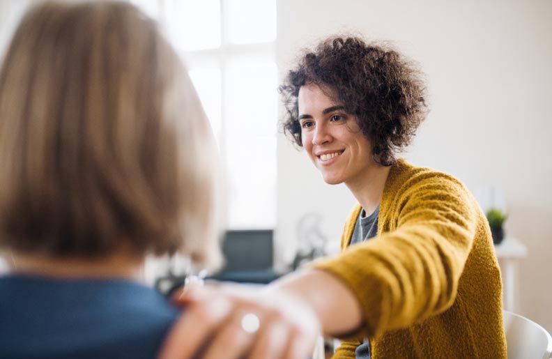 TraumaSolutions A woman with curly hair in a yellow sweater smiles and places a comforting hand on another person's shoulder, offering gentle support amid loss or trauma.