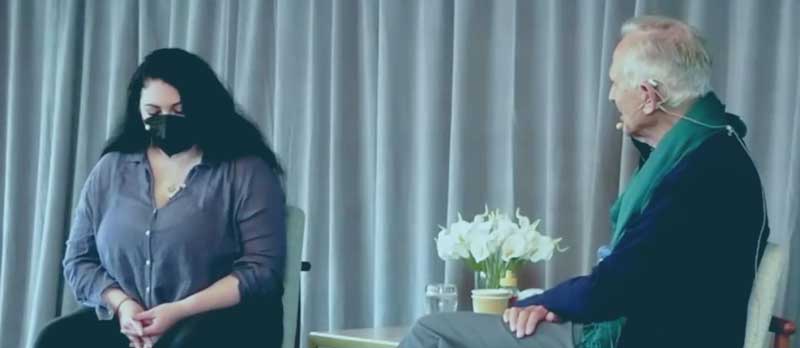 TraumaSolutions A woman wearing a mask sits with hands clasped, facing an older man with a microphone. They are seated indoors with a table of flowers and water between them, engaged in a thoughtful conversation about somatic healing and restoring resilience after complex trauma.