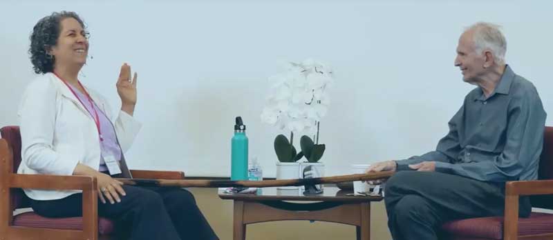 TraumaSolutions Two people sit facing each other in chairs, talking and smiling, as they share stories of resilience. A small table holding a white orchid and water bottles between them adds a calming touch to their moment of connection.