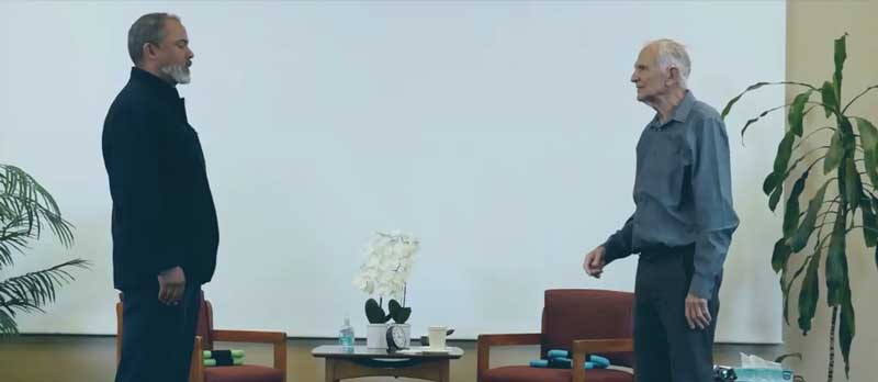 TraumaSolutions Two men stand facing each other in a room with chairs, a potted orchid, and large plants, creating an atmosphere conducive to somatic healing. A white screen is in the background, setting the scene for conversations about restoring resilience after complex trauma.