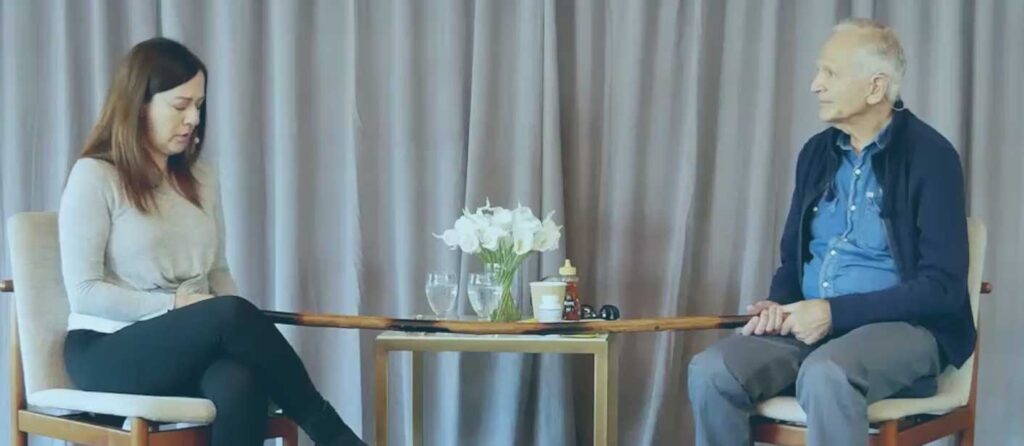TraumaSolutions Two people sit facing each other on chairs with a table between them holding flowers, glasses of water, and other items; a curtain hangs in the background, creating a calming space conducive to somatic healing and restoring resilience after complex trauma.