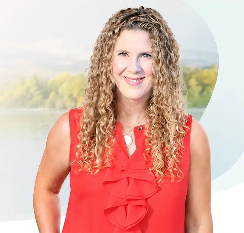 TraumaSolutions Smiling woman with long curly blonde hair wearing a sleeveless red blouse, standing outdoors with a blurred background of trees and water, embodying the search for connection amidst nature’s serene beauty.