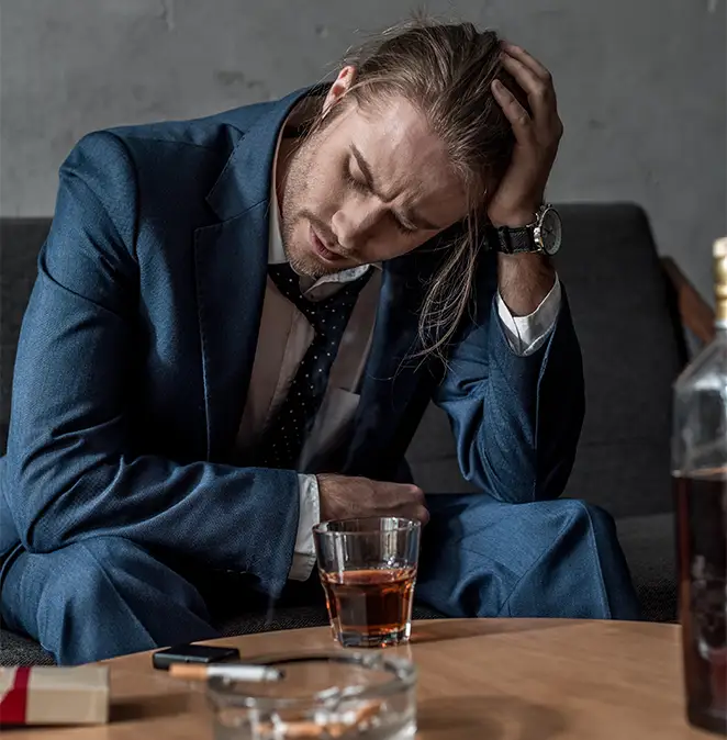 TraumaSolutions A man in a blue suit sits on a couch with his head in his hand, looking distressed. A glass of whiskey, a pack of cigarettes, and a bottle are on the table in front of him—silent witnesses to his Search for Connection amid loneliness.