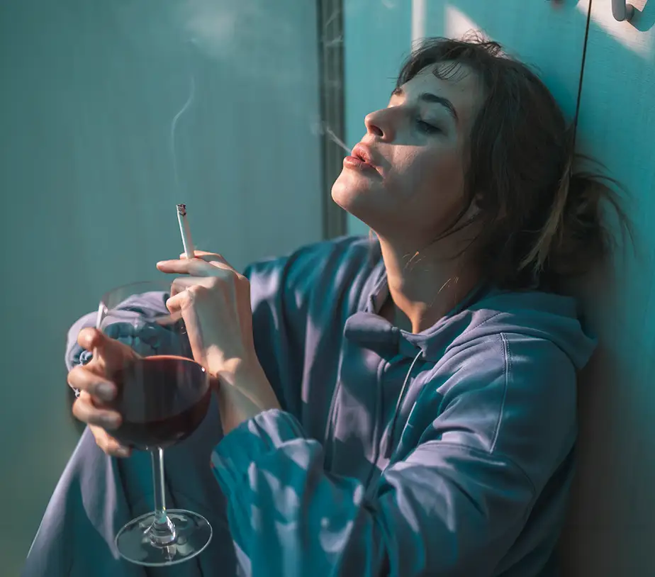 TraumaSolutions A woman in a blue hoodie sits on the floor against a wall, holding a glass of red wine in one hand and a lit cigarette in the other, lost in quiet contemplation and the search for connection.