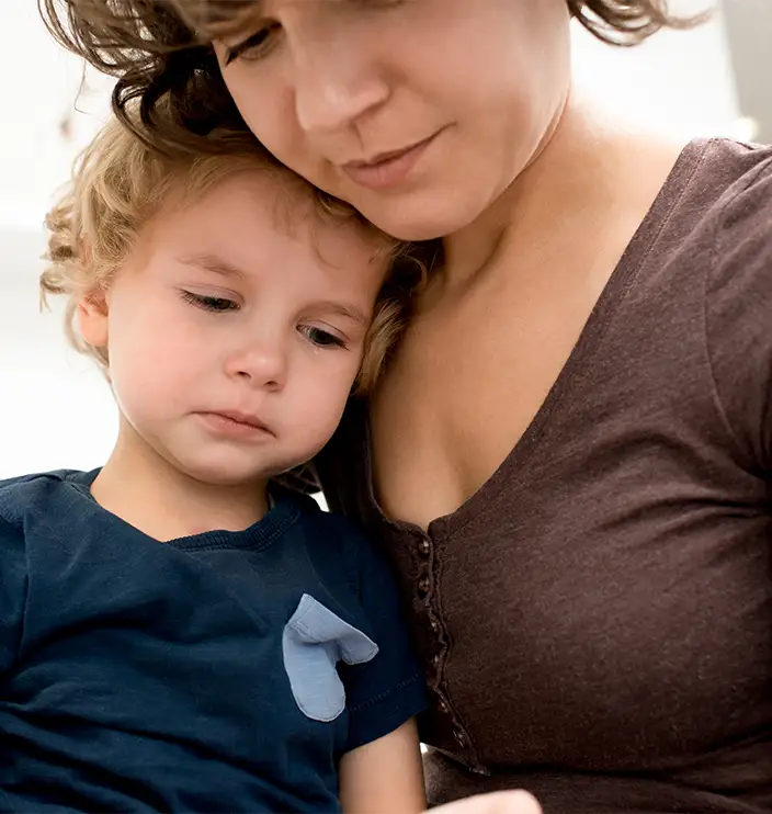 TraumaSolutions Woman holds and comforts a sad young child who is resting their head on her chest, illustrating a quiet search for connection. Both have light skin and brown hair. The child is wearing a dark blue shirt with a light blue patch.