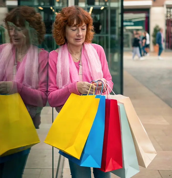 TraumaSolutions A woman with curly red hair stands outside a glass storefront holding several colorful shopping bags in both hands, her gaze suggesting a search for connection as she scans the bustling street.