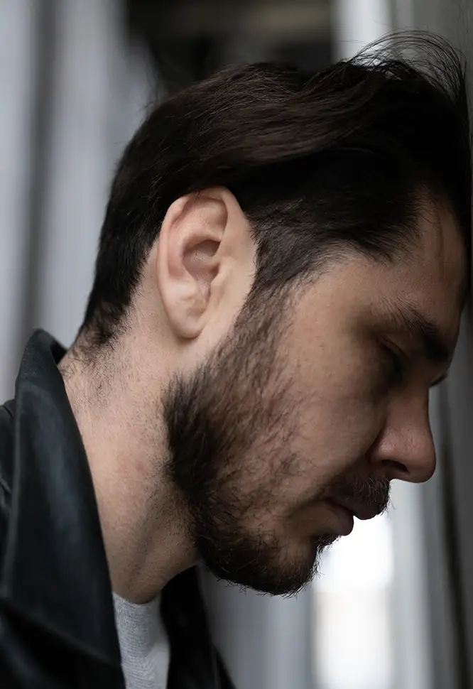 TraumaSolutions Man with dark hair and beard leans his forehead against a wall, eyes closed, wearing a dark jacket, in a close-up side profile view—a quiet moment that suggests a search for connection.