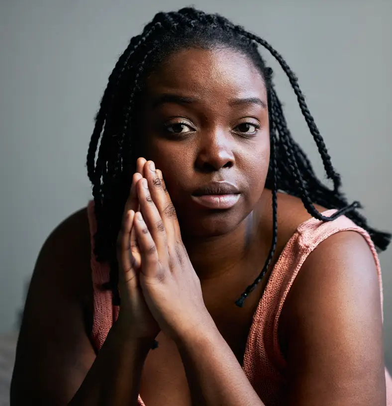 TraumaSolutions A person with braided hair sits indoors, hands pressed together near their face, looking directly at the camera with a neutral expression, as if quietly engaged in a search for connection.