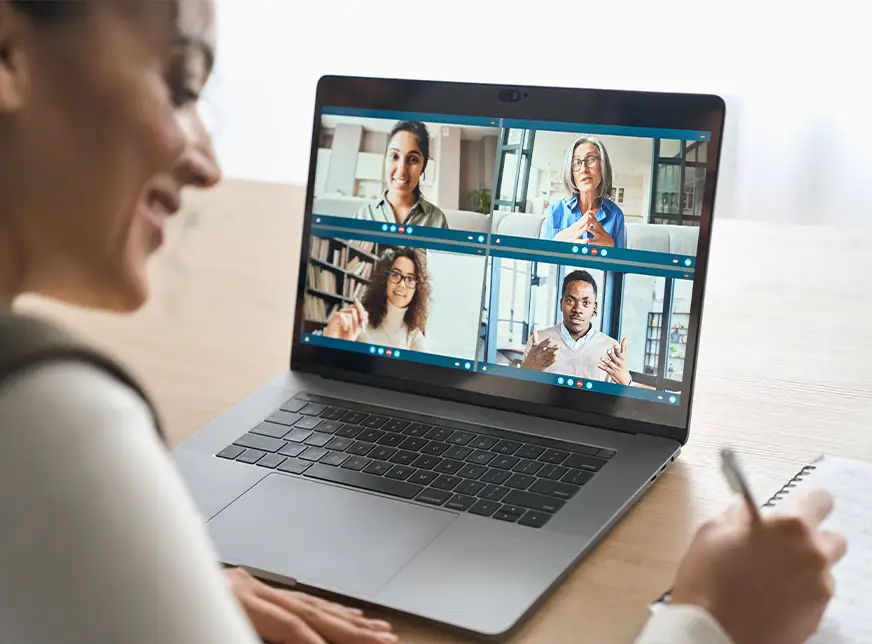 TraumaSolutions Person taking notes while participating in a video conference call on a laptop with four people displayed on the screen, illustrating the ongoing search for connection in today’s digital workspace.