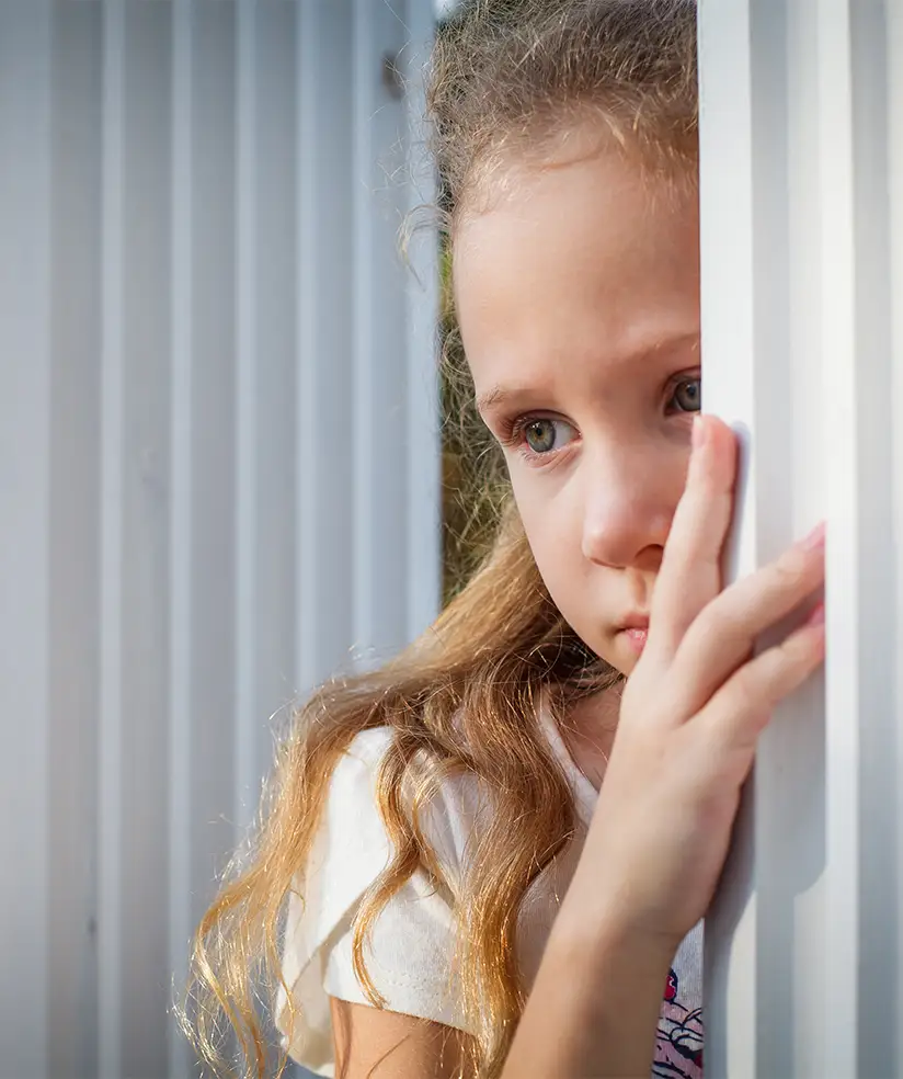 TraumaSolutions Young girl with long hair stands by a white fence, looking thoughtful and holding onto the fence with one hand, as if lost in a quiet search for connection.