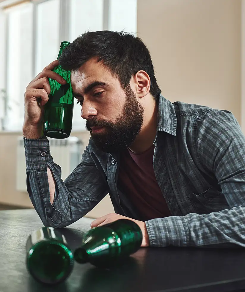 TraumaSolutions A man with a beard sits at a table holding an empty green bottle to his head, with two more empty bottles lying in front of him—a quiet moment reflecting his search for connection.