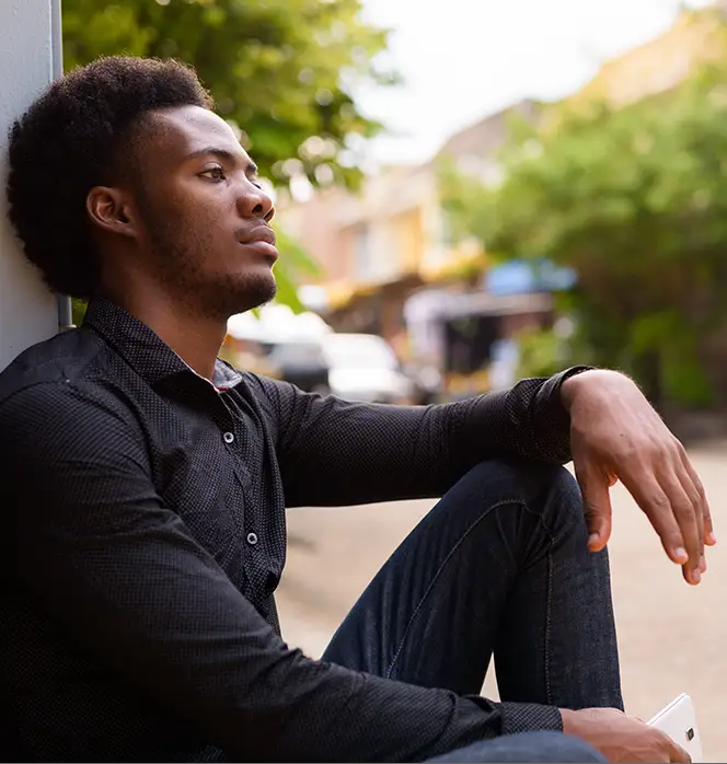 TraumaSolutions A man in a black shirt and jeans sits outdoors against a wall, looking thoughtful, as if deep in search of connection, with greenery and blurred buildings in the background.