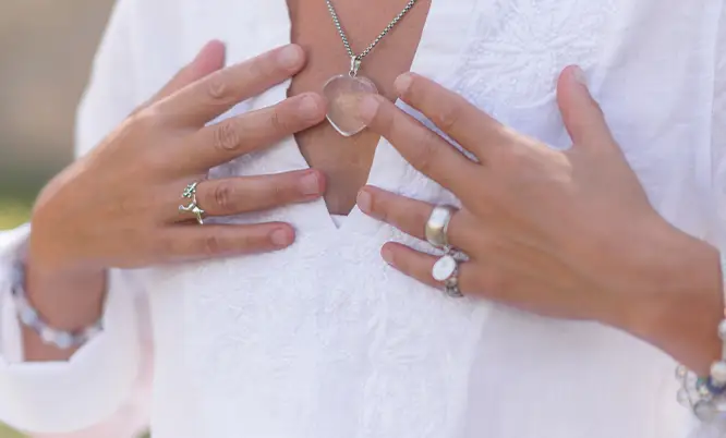 TraumaSolutions Person wearing a white shirt holds a heart-shaped pendant necklace, subtly expressing a search for connection while showing rings and bracelets on both hands.